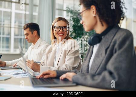 Smiling woman manager working on laptop in modern coworking while sitting near colleagues Stock Photo