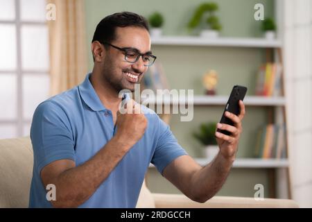 Excited indian man celebrating by seeing mobile phone while sitting on sofa at home - concept of good news, job promotion and successful Stock Photo