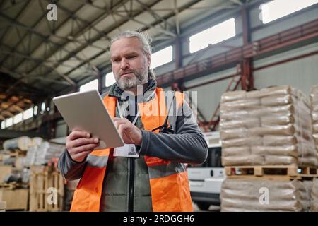 Mature foreman in reflective clothing using tablet pc to control the number of cargo in warehouse Stock Photo