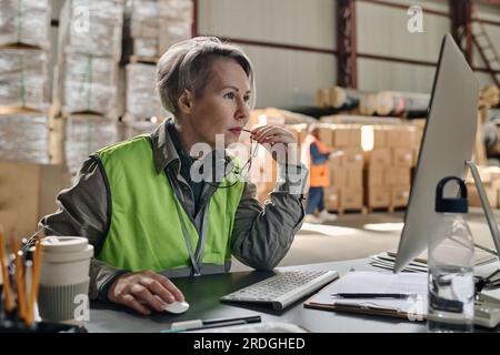 Serious foreman sitting at table in front of monitor and checking online documents on computer Stock Photo