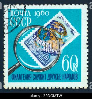 A postage stamp issued in the USSR in 1960 for the International Collectors' Day. The turquoise stamp features a postage stamp under a magnifying glass. The smaller stamp within a stamp features the text: 'защита мира есть дело всех народов мира' 'Defending peace in the world is all nations' work'. The text 'Филателия служит дружбе народов' – 'Philately serves the friendship of peoples' is printed below. Face value: 60K (60 kopeks). Stock Photo