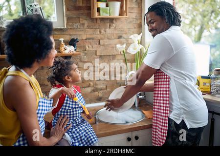 Cute little girl blowing bubbles in the kitchen by the sink, while her father is washing dishes. Cheerful family together. Home, family, lifestyle con Stock Photo