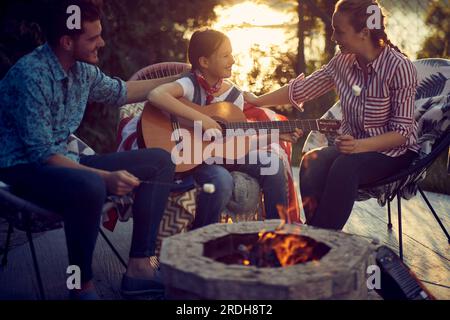 Young girl playing guitar outdoors by the campfire, mother and father singing along. Lovely family together. Togetherness concept. Stock Photo