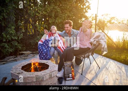 Joyful man and woman with daughter sitting by fireplace outdoors, roasting marshmallows and laughing, having a good time together. Home, family, lifes Stock Photo