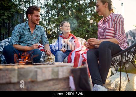 Joyful and happy family of man, woman and girl sitting outdoors by a fireplace roasting marshmallows enjoying sunny summer day together. Family, holid Stock Photo