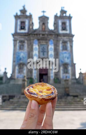 holding Pastel de nata in Porto Portugal in front of beautiful white blue Church of Saint Ildefonso with azulejo tiles . Stock Photo