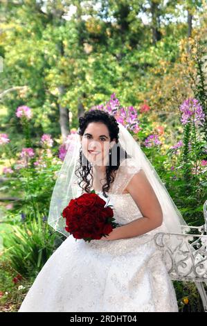 Beautiful bridge sits surrounded by flowers and garden.  She is holding a bouquet of red roses and smiles happily. Stock Photo