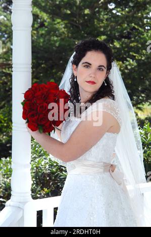 Beautiful bride holds a bouquet of red roses.  She is standing in a white gazebo and looks dreamy and romantic. Stock Photo