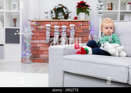 A tiny boy sits in a room on a gray sofa holding a cuddly toy Stock Photo
