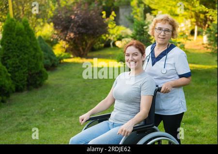 An elderly female nurse walks with a middle-aged woman in a wheelchair in the park.  Stock Photo