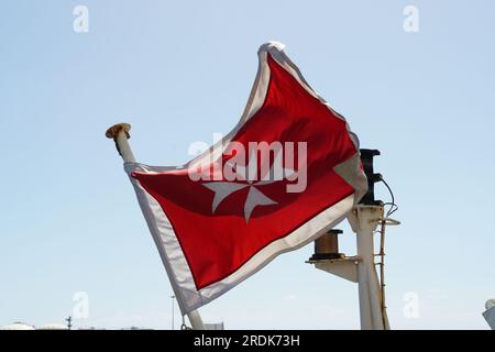 Merchant flag of Malta, red banner with white Maltese cross, flying in aft part oft merchant container vessel in Cape Town port. Stock Photo