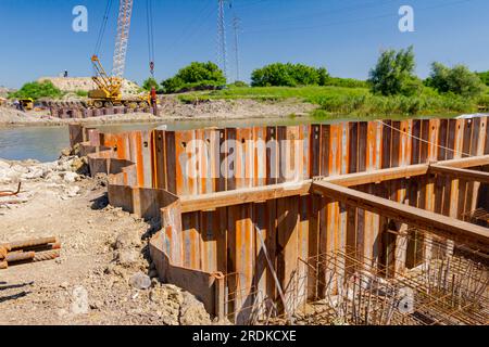 Installed rusty metal piles along river bank built in for bridge foundation at the construction site. Across the water is yellow obsolete mobile crane Stock Photo