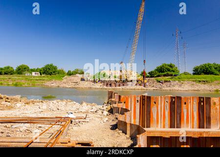 Installed rusty metal piles along river bank built in for bridge foundation at the construction site. Across the water is yellow obsolete mobile crane Stock Photo