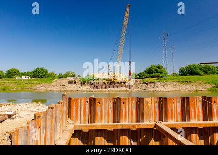 Installed rusty metal piles along river bank built in for bridge foundation at the construction site. Across the water is yellow obsolete mobile crane Stock Photo