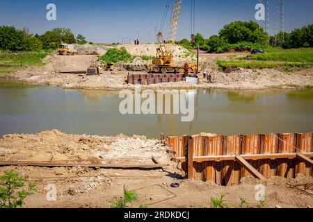 Installed metal piles along river bank built in for bridge foundation at the construction site. Across the water is yellow obsolete mobile crane with Stock Photo