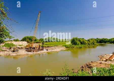 View through across the water is yellow obsolete mobile crane with pneumatic hammer. Installed metal piles along river bank built in for bridge founda Stock Photo