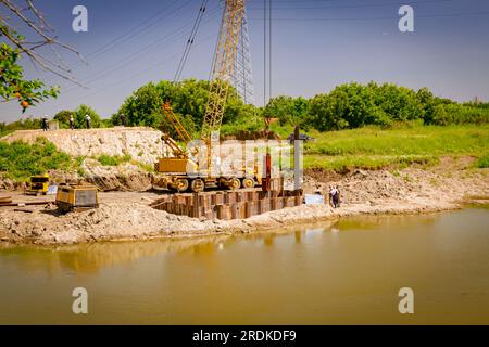 View through across the water is yellow obsolete mobile crane with pneumatic hammer. Installed metal piles along river bank built in for bridge founda Stock Photo