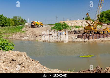 View through across the water is yellow obsolete mobile crane with pneumatic hammer. Installed metal piles along river bank built in for bridge founda Stock Photo