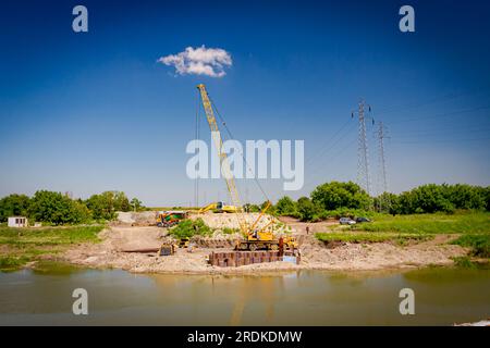 Across the water is yellow obsolete mobile crane with pneumatic hammer. Installed metal piles along river bank built in for bridge foundation at the c Stock Photo