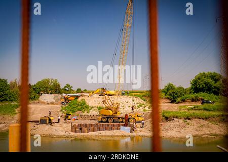 View through across the water is yellow obsolete mobile crane with pneumatic hammer. Installed metal piles along river bank built in for bridge founda Stock Photo