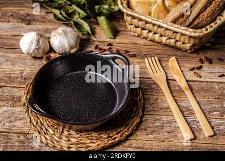 Empty black deep plate on a rustic wooden table with cutlery, bread, garlic, cayenne pepper and bay leaf. Stock Photo