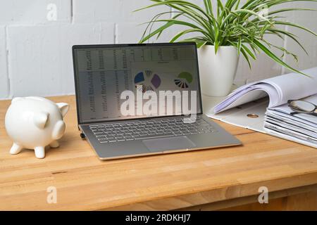 Wooden desk with laptop and number charts on the screen, piggy bank and file folder in front of a white painted wall in a home office, concept for bus Stock Photo