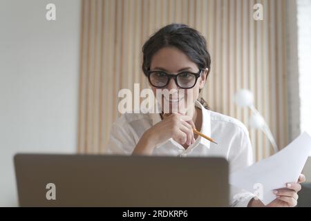 Young smiling italian business woman wearing glasses using laptop at work, happy female employee looking at screen with smile, reading email with good Stock Photo