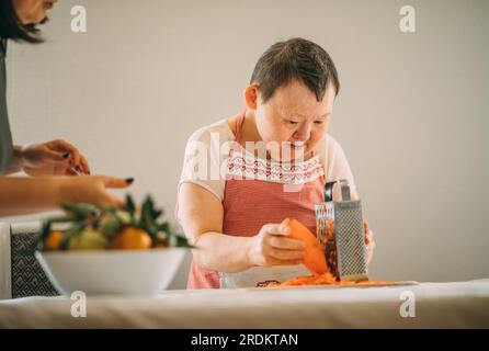 elderly woman with Down syndrome gratefully learns to grate carrots with the guidance of a teacher Stock Photo