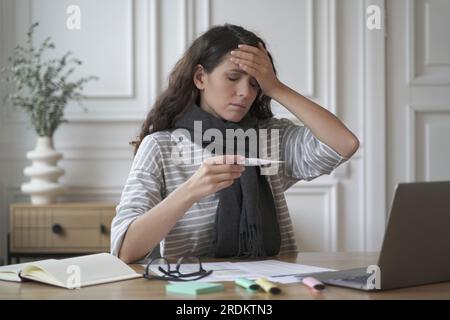Sick young woman freelancer sitting at workplace and touching forehead while taking body temperature, sick hispanic female employee reading high fever Stock Photo