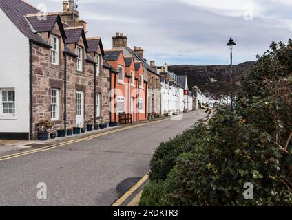 View eastwards along West Shore Street, Ullapool with sunlit colourful buildings under cloudy sky and gulls flying above Stock Photo