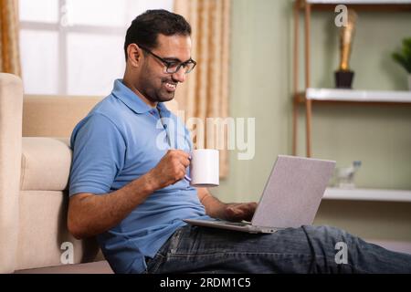 Relaxed Indian man using laptop by drinking coffee while sitting on sofa at home - concept of Remote work, fortable workspace and Digital lifestyle Stock Photo
