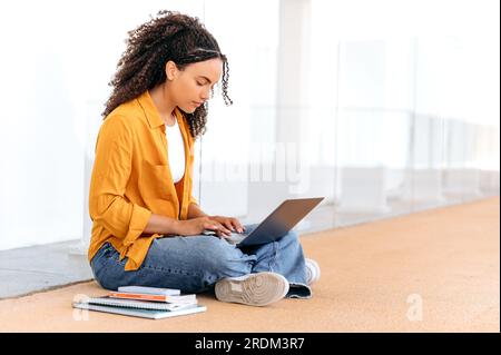 Focused busy lovely stylish curly haired brazilian or hispanic female student, sit near the university campus, using laptop, preparing for the exam, doing homework, project task, working on a project Stock Photo