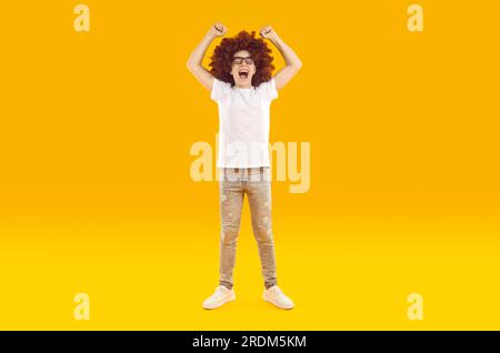 Funny boy in red curly wig screaming with joy with open mouth and with his hands up in fist. Stock Photo