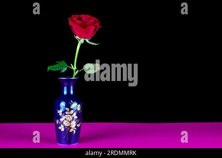 Blue decorated flower vase with single red rose on a black background and pink base Stock Photo