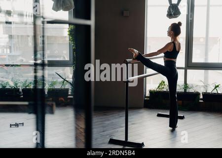 Slim red-haired ballerina stands in split position beside ballet barre in gym, light window background. Indoor sport and stretching showcased beautifu Stock Photo