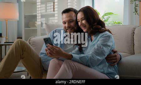 Millennial couple Caucasian man woman married husband wife at home talking using smartphone together happy family discuss offers choosing shopping Stock Photo