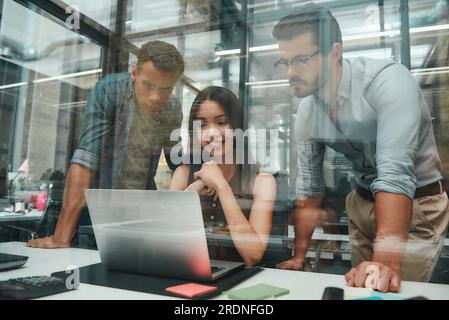 Busy day. Group of three young employees looking at screen of laptop and discussing new ideas while working in modern open space Stock Photo