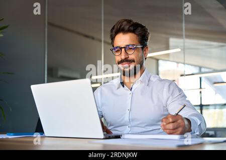 Busy smiling young Latin business man working in office using laptop writing. Stock Photo