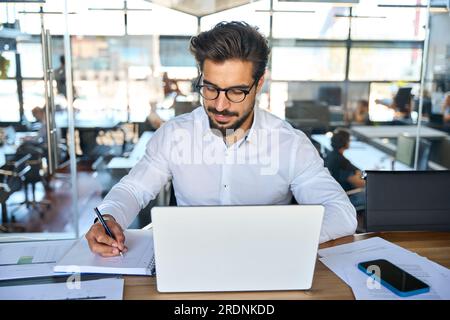Busy young Latin business man working in office on laptop writing notes. Stock Photo