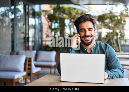Smiling Latin business man talking on cellphone working on laptop outdoors. Stock Photo