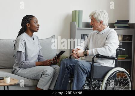 Side view portrait of black young woman as nurse helping senior in retirement home and using digital tablet Stock Photo
