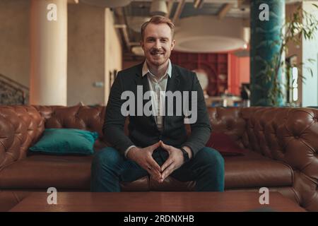 Smiling businessman sitting on sofa and looking at camera during working day in modern coworking Stock Photo