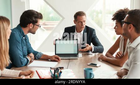 Analyzing results. Serious mature man in formal wear showing something on the digital tablet and discussing it with his multicultural team while Stock Photo