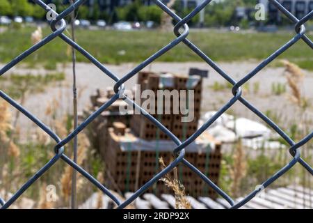 Chain link fence in front of orange bricks on wooden pallets - grassy field and cars in background - clear blue sky Stock Photo