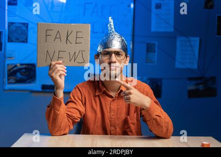 Male conspiracy theorist in a protective foil cap and glasses debunks myths holding poster fake news. Conspiracy theory concept. The schizophrenic Stock Photo