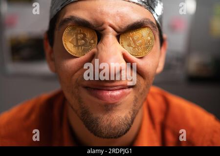 Strange stupid man conspiracy theorist in protective foil cap holds two bitcoins in his eyes. Male with bitcoins in eyes. Bitcoin is a cryptocurrency Stock Photo