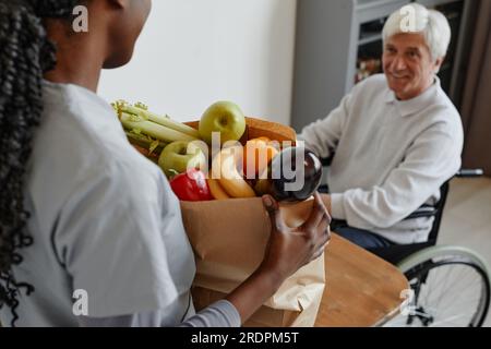 Closeup of nurse bringing bag with groceries to smiling senior man in wheelchair Stock Photo