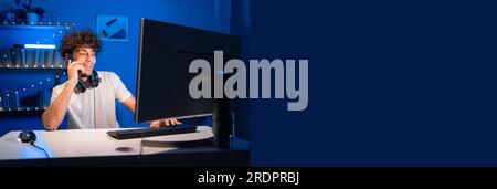 A Handsome Arabic guy gamer in a headset at a computer calls on the phone with a smile on his face. Stock Photo