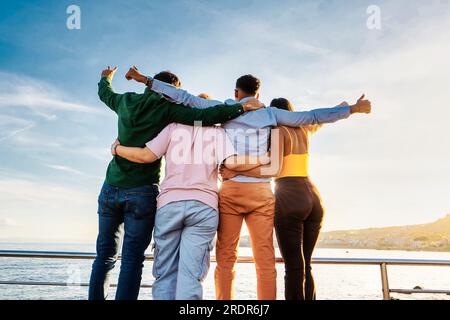 Four friends of diverse ethnicities, two men and two women, are seen from behind, embracing and facing the setting sun. Stock Photo