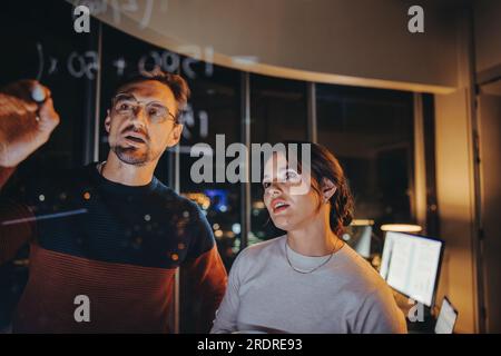 Team of business professionals collaborating in a late-night brainstorming session. Two business people discussing and analysing ideas in a glass-wall Stock Photo